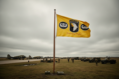 A yellow military flag with the words 'Airborne' and 'Reserve Infantry' and three circular emblems, flying on a flagpole in an open field with vintage aircraft and soldiers in the background under a cloudy sky. WWII airfield